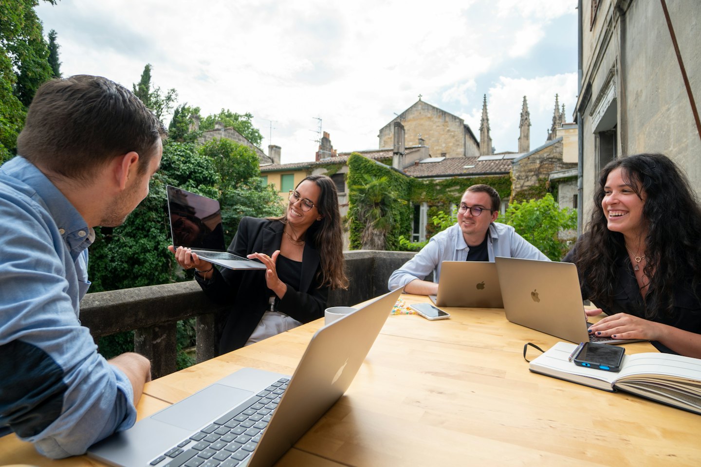 Vier junge Berufstätige arbeiten draußen an einem Tisch mit Laptops zusammen.